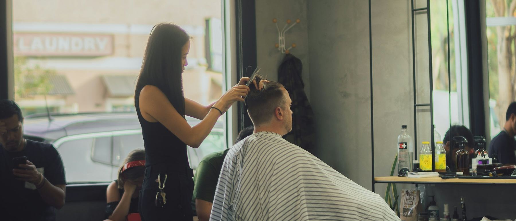 A stylist cutting a client's hair in a small neighbourhood shop, with customers waiting.